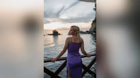 EmilyGiffin poses on a wooden deck overlooking the ocean, wearing a purple slip dress with a high slit and her blonde hair down.