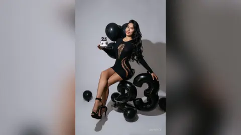Smiling woman in a black cocktail dress holding a 23 birthday sign with black balloons in a studio setting.