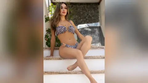 LinaVon sits on white stairs wearing a leopard print bikini, posing with one leg bent and her hands resting on the steps.