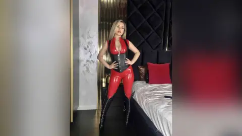 Blonde woman in a red latex bodysuit and black boots posing in a modern luxury bedroom.