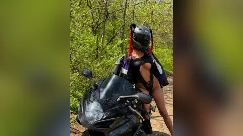 Woman with red braids in a black bodysuit sitting on a black sport motorcycle in a lush green forest.