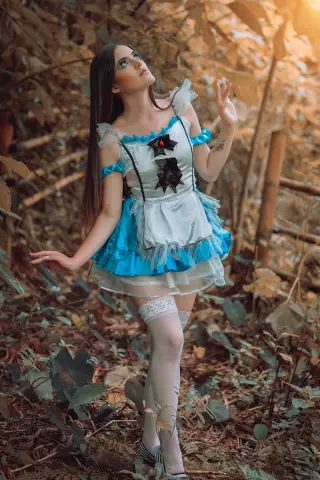 AlicceGray poses in a blue and white Alice in Wonderland style dress and white thigh-high stockings while standing in a leafy forest setting.