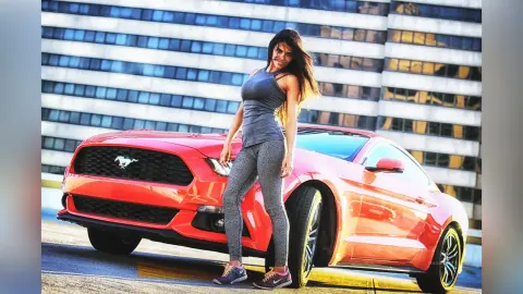 DrellaWolf — Stylish woman in grey athletic wear posing confidently next to a red Ford Mustang in a modern city setting.