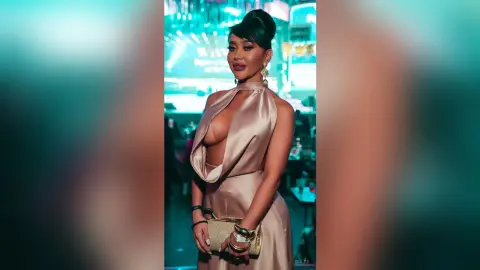 Dolce poses in a champagne satin dress with a deep plunging neckline, wearing gold earrings and holding a gold clutch.
