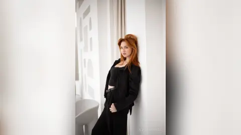AlbertaCorrell is a redhead wearing a black crop top and black pants, posing against a white wall.