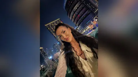 Smiling woman in a white lace blouse posing against a night cityscape with glowing skyscrapers.