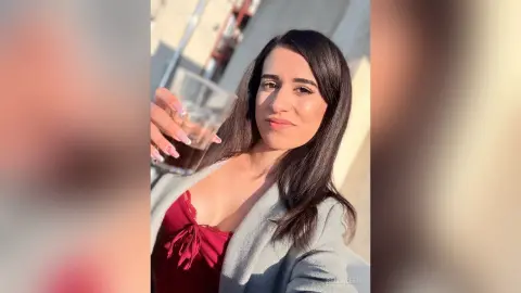 Smiling woman in a red lace camisole and grey cardigan enjoying a drink outdoors in warm sunlight.