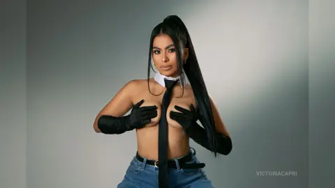 Young woman in black gloves and tie with denim jeans posing against a grey studio background.