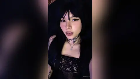 Young woman with black hair and butterfly neck tattoo wearing a black lace top in moody low lighting.