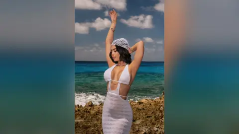 Young woman in a white crochet maxi dress posing on a rocky beach under a blue sky.