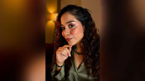 Smiling woman with wavy hair wearing an olive green blouse and beaded necklace in warm indoor lighting.