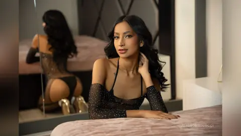 AlissonBelucci poses in a black sheer mesh bodysuit with white polka dots, showing her cleavage and back while leaning on a pink bed.