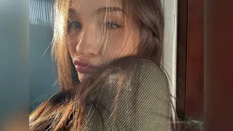 Young woman with long brown hair wearing a green ribbed sweater in warm, sunlit indoor lighting.