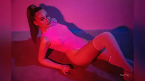 BeaNix lies on a pink floor wearing a bright pink sleeveless bodysuit and white thigh-high stockings.