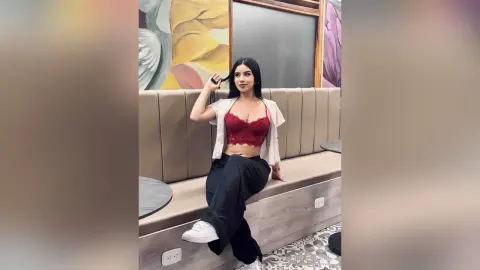 Young woman in a red lace crop top and black trousers sitting in a modern cafe setting.