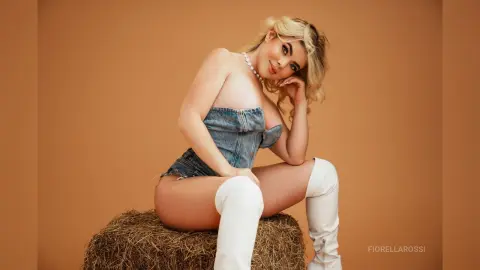 FiorellaRossi poses naked in denim shorts and white thigh-high boots while sitting on a hay bale.