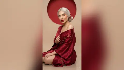 Elegant woman with silver hair in a red silk robe posing against a minimalist red and beige background.