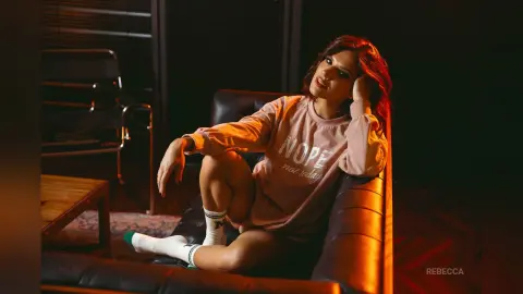 Rebecca sits on a black leather couch wearing a pink sweatshirt and white socks, looking at the camera in warm lighting.