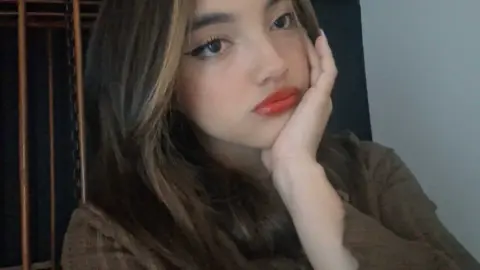 Young woman with long brown hair and red lipstick posing in a cozy brown sweater for a moody selfie.
