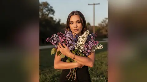 Beautiful woman in a black dress holding a large bouquet of purple lilacs in a soft outdoor sunset setting.