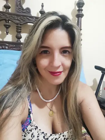 Smiling woman with blonde highlights wearing a leopard print top and gold necklace in a cozy bedroom setting.