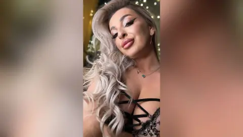 Glamorous woman with long silver hair and winged eyeliner wearing a black lace corset in a festive setting.