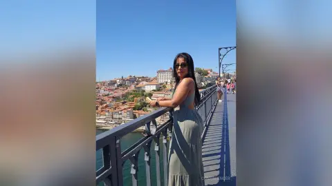 Stylish woman in a sage green maxi dress leaning on a bridge railing overlooking a coastal European town.