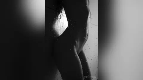 RubyAttwood poses naked in a side profile view, showing her bare chest and stomach against a light background with water droplets visible on her skin.