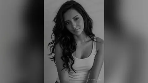Portrait of a smiling young woman with long wavy hair wearing a white tank top in a moody black and white setting.