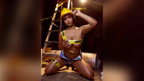 MiaKorz poses in a yellow hard hat and denim shorts, wearing yellow danger tape across her bare breasts and body.