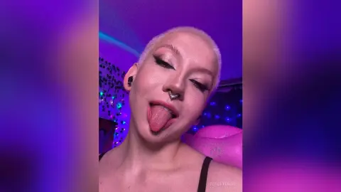 JoyceTreier with short blonde hair and nose ring sticks her tongue out at the camera in a room with purple lighting.