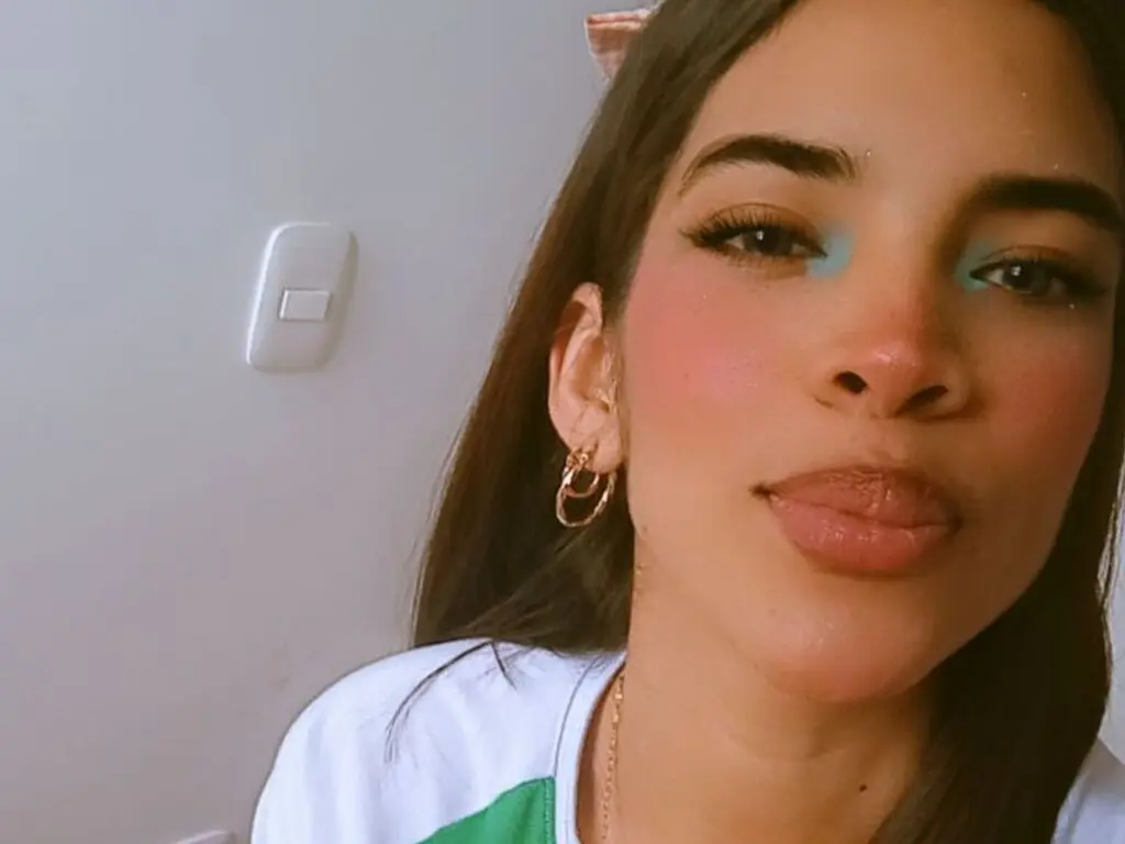 MeliSanders looks into the camera with blue eyeshadow and gold hoop earrings while wearing a white shirt.