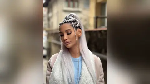 Woman with long silver braided hair and light blue top posing outdoors in a stylish urban setting.