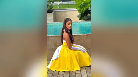 TefyRusso wearing a bright yellow ballgown and white gloves, sitting by a blue swimming pool with her long dark hair in a high ponytail.