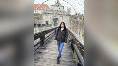 Young woman in a black puffer jacket and jeans walking on a wooden bridge under festive fairy lights.