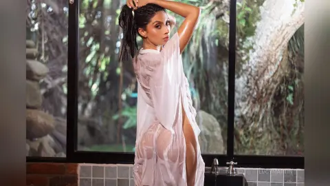 LucyMartin poses in a sheer white robe with a high slit, showing her bare legs against a background of trees and glass.