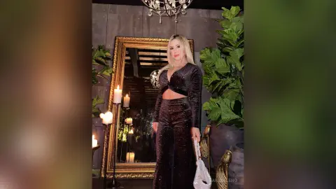 SaraBeleno stands in front of a gold mirror wearing a black long-sleeved crop top and matching high-waisted trousers.