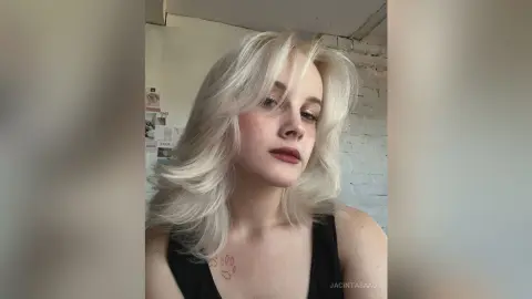 Young woman with platinum blonde wavy hair wearing a black tank top in a bright indoor setting.