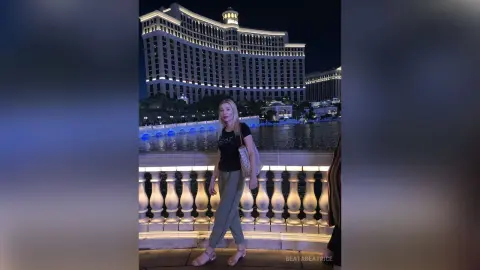 Blonde woman in a black t-shirt and grey trousers standing by a lit fountain at a luxury night resort.