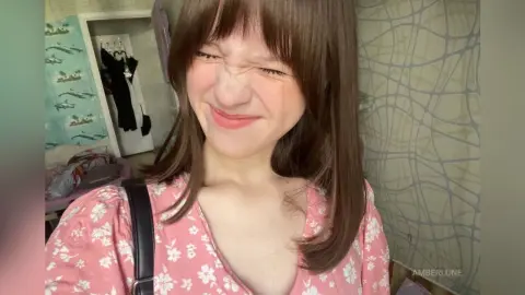 AmberLune smiles with her eyes squinted, wearing a pink floral v-neck top with her long brown hair and bangs framing her face.