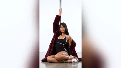 Stylish woman in a black bodysuit and oversized burgundy faux fur coat posing against a white studio background.