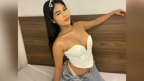 MadeleinSinclair poses in a white corset top and denim shorts, leaning back against a wooden headboard.