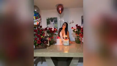 Smiling woman in a white dress celebrating a birthday with cake, roses, and balloons at a festive indoor party.