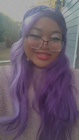Smiling woman with long purple hair and glasses wearing a cozy beige sweater in a soft outdoor setting.