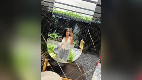 Young woman in a white sleeveless top and grey trousers posing in a modern marble bathroom mirror.