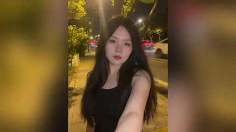 Young woman in a black tank top smiling on a city sidewalk at night under warm streetlights.