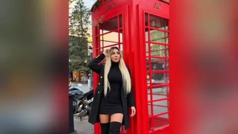 Stylish woman in a black dress and long coat posing by a classic red London telephone booth on a city street.