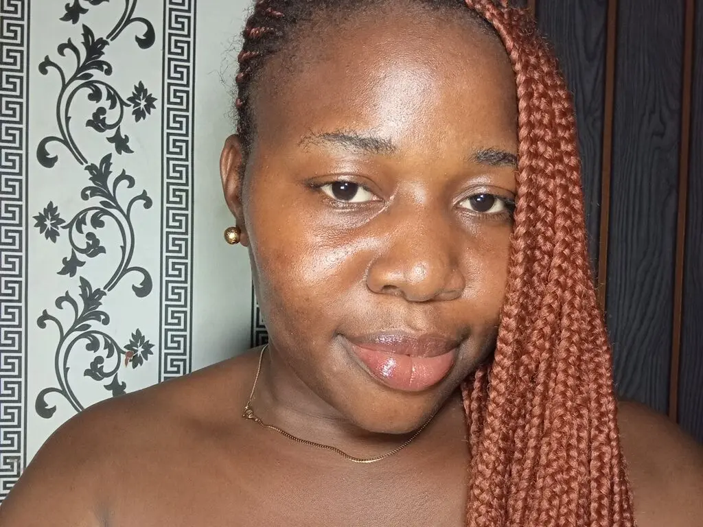 Smiling Black woman with long reddish-brown braids and gold earrings posing in a room with patterned wallpaper.