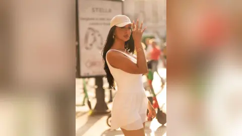 NicoleBella wears a white tank top and matching white skirt with a white cap, posing outdoors in the sunlight.