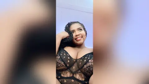 Smiling woman with long braids wearing a black sheer lace bodysuit in a bright indoor setting.